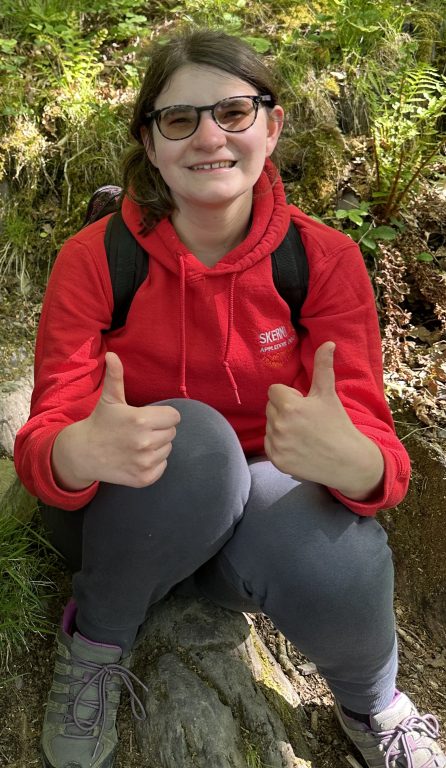 Student smiling sat on a rock wearing a red hoodie and walking boots doing a thumbs up