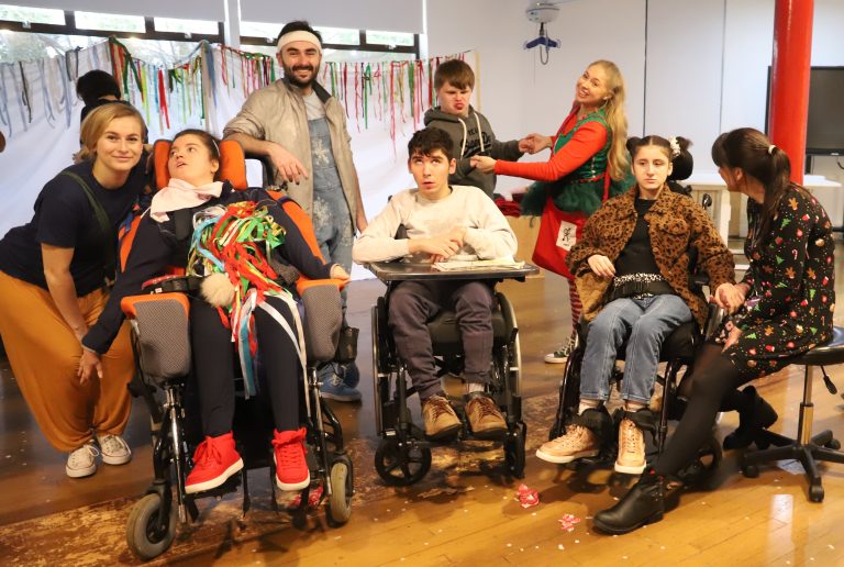 Three students in wheelchairs and one dancing in the back with smiling theatre group members.