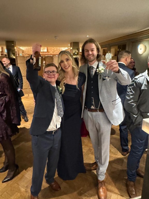 Three people in smart wedding gear holding up beer bottle and champagne glass in a 'cheers' action.