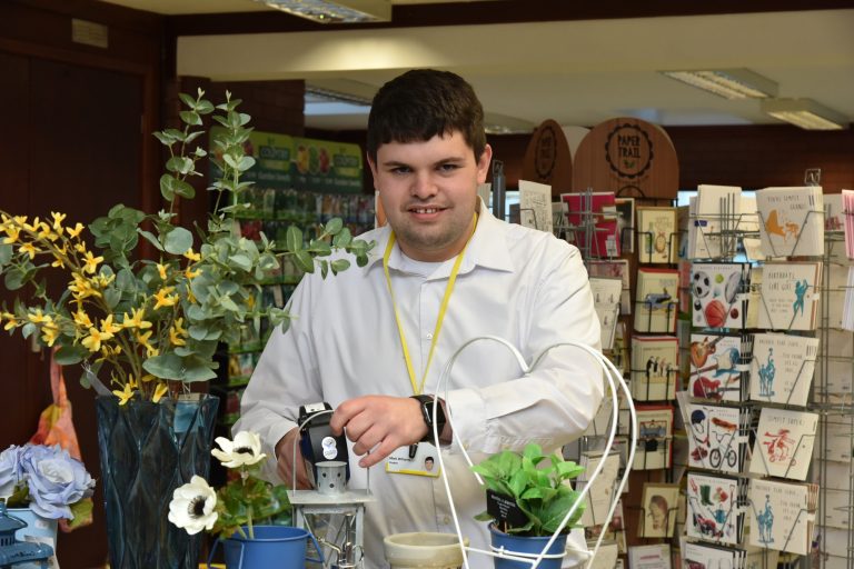 Smiling student wearing smart white shirt, surrounded by greeting cards and flowers in the Garden Centre shop, and holding a pricing gun.