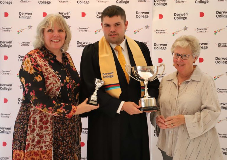 Student Mark wearing a smart suit, black gown and gold graduation sash holding a large and a small silver trophy. Pictured with smiling College Principal and Chair of Governors.