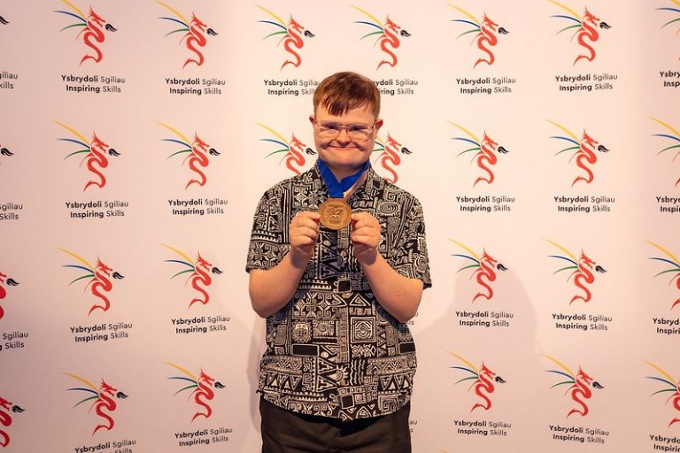 Student holding a large Bronze medal around his neck in front of a background with Inspiring Skills logos.