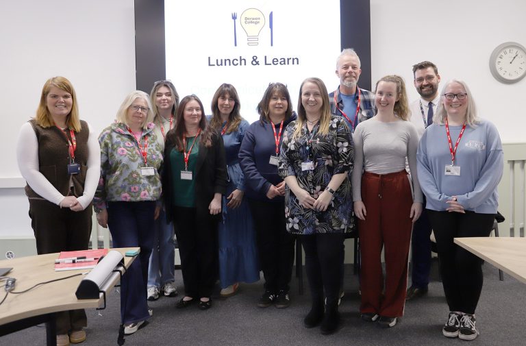 Group of employers and staff stood in front of a white board that says Lunch & Learn on it along with a logo with a lightbulb, knife and fork.