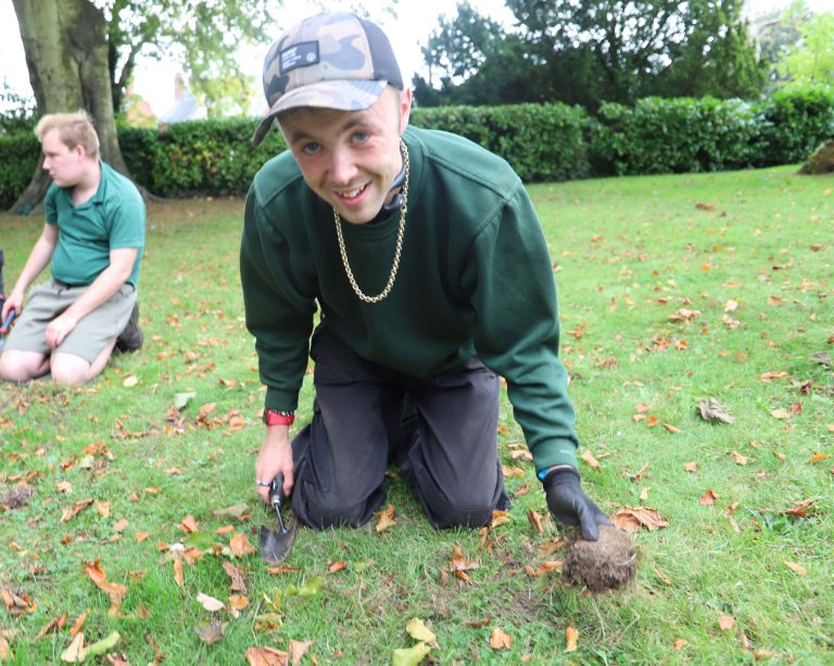 Student in green work sweatshirt and cap kneels on the grass holding a trowel and a divot of grass.